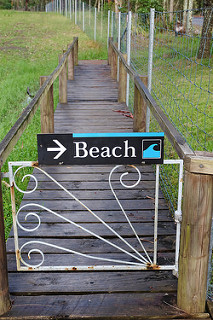 Sign on a small gate says "Beach", with an arrow. Beyond the gate is a wooden walkway into the distance.