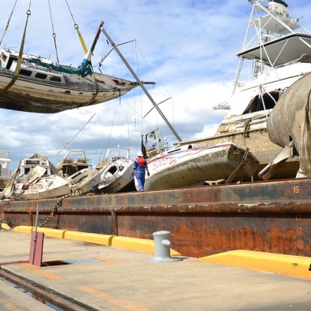 A crane moving a yacht onto a pier, where there are many other boats that have been wrecked in a hurricane.